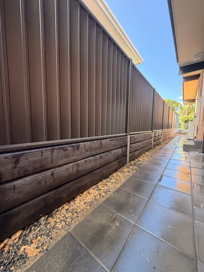 Dark woodgrain sleeper retaining wall beneath a Colorbond fence beside a tiled path